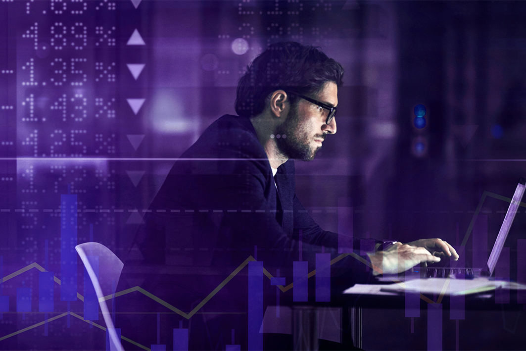 At night, man sitting at a desk, typing at laptop. Abstract overlay images of share prices going up and down.